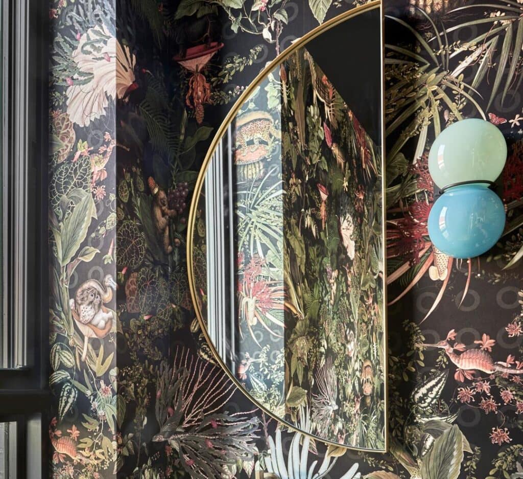 Decorative mirror with gold frame reflecting tropical wallpaper and two pastel glass orbs.